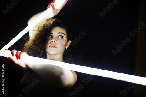 Curly brunette plus size model with two long led lamp at black room. photo