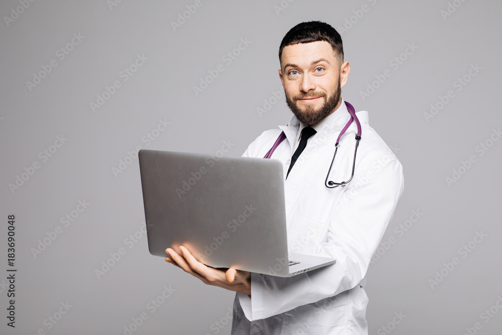 Portrait of a handsome young male doctor with stethoscope dressed in uniform holding laptop computer while standing and looking at camera isolated over white background Portrait of a handsome young male doctor with stethoscope dressed in uniform holding laptop computer while standing and looking at camera isolated over white background