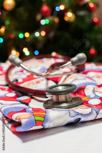 Close up of a red stethoscope on Christmas Santa Claus scrubs on a white background with blurred Christmas tree with colorful lights in the background. Christmas in hospital or healthcare concept