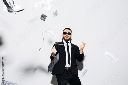 Handsome man in suit and sunglasses pointed up on fluy cash money on white background. Businessman standing in the rain of money.