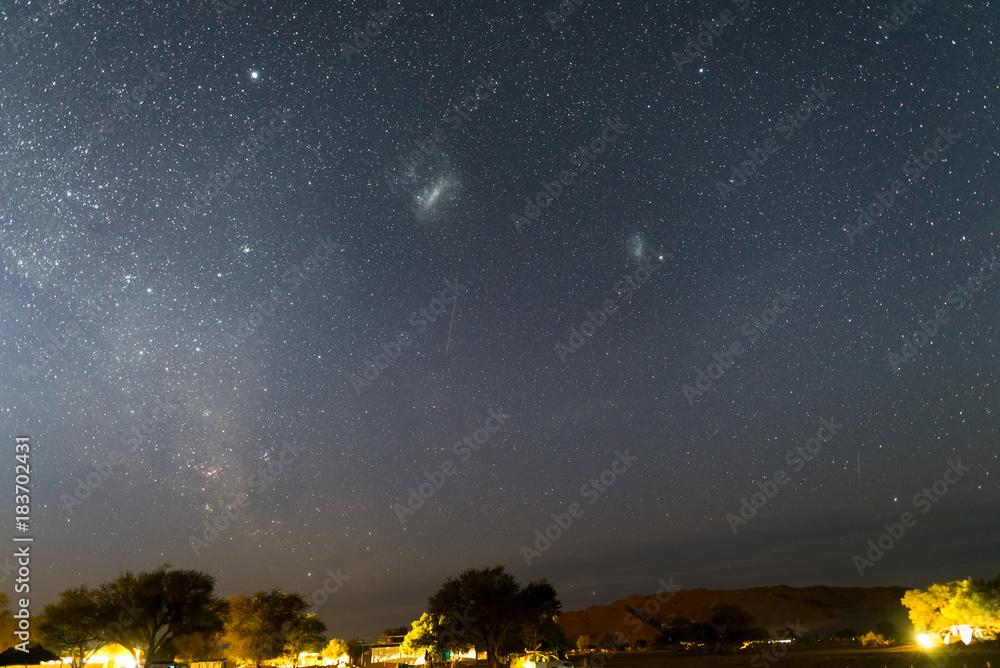 Fototapeta premium The starry sky and the majestic Magellanic Clouds, outstandingly bright, captured in Africa. Acacia trees and straw hut in the foreground. Adventure into the wild.
