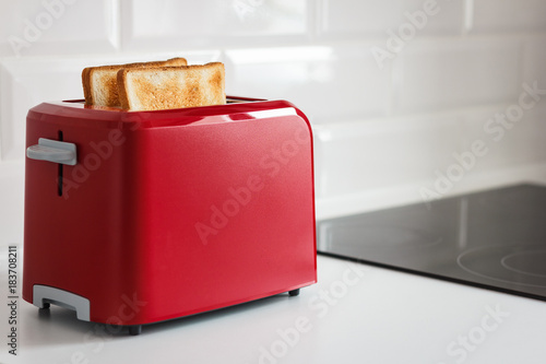 Red toaster with toasted bread for breakfast inside. White background. White kitchen table.