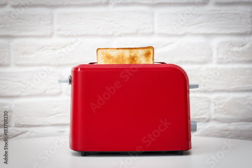 Red toaster with toasted bread for breakfast inside. White table. White brick wall.