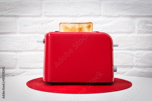 Red toaster with toasted bread for breakfast inside. Red table napkin. White table. White brick wall.