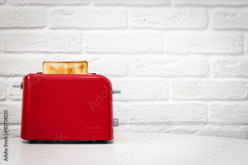 Red toaster with toasted bread for breakfast inside. White table. White brick wall.