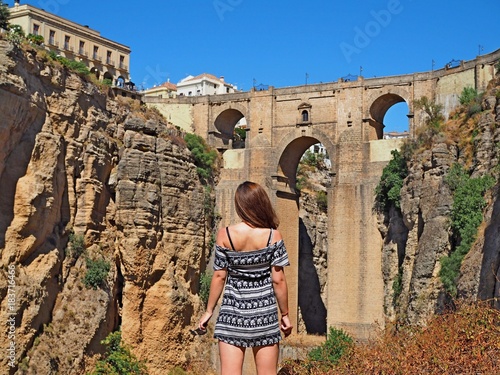 Girl in a dress and long hair standing turned away from the camera and looking at the ancient bridge in Ronda Spain, around mountains and stones