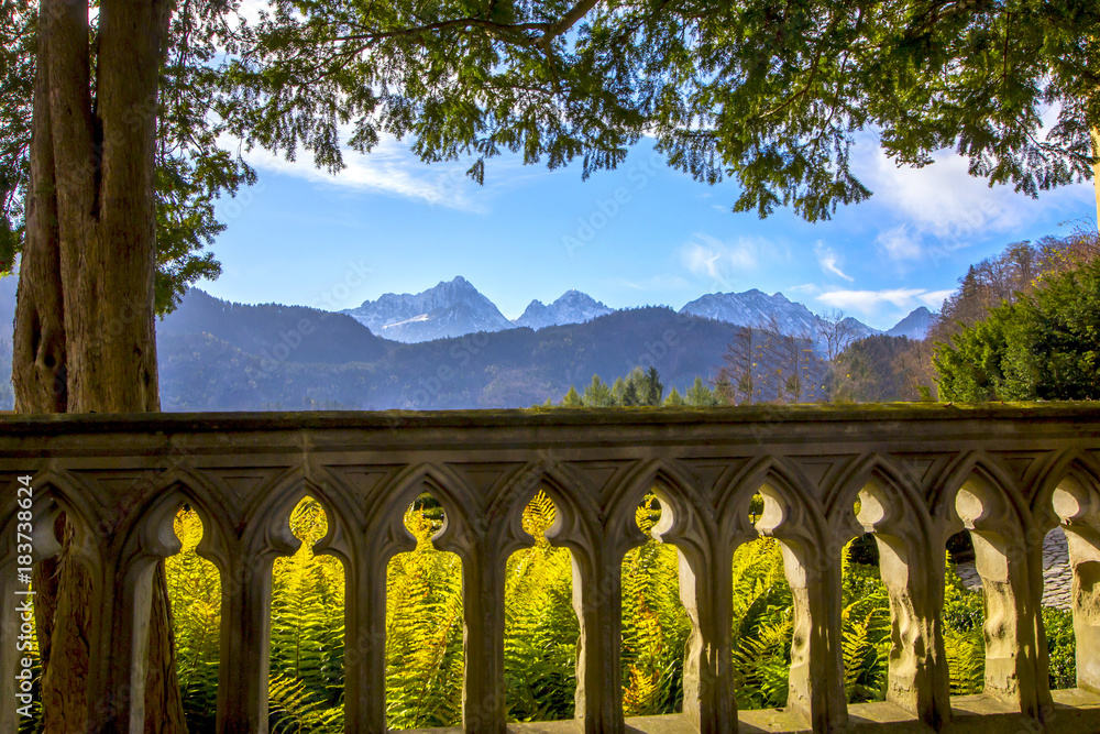 View of the Mountain Alps from the terrace with a beautiful fence and coniferous trees View of the Mountain Alps from the terrace with a beautiful fence and coniferous trees