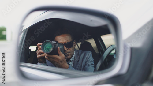 Reflection in side mirror of Paparazzi man sitting inside car and photographing with dslr camera