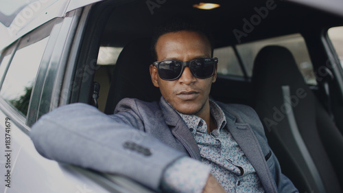 Portrait of serious businessman in sunglasses sitting inside car and looking into camera outdoors