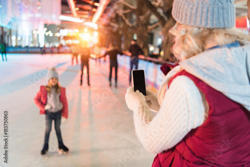 young woman with smartphone photographing cute little daughter on skating rink