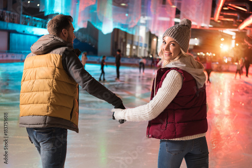 back view of happy young couple holding hands and skating on rink