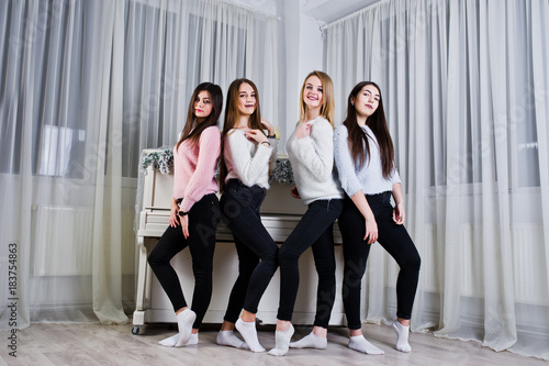 Four cute friends girls wear on warm sweaters and black pants against old piano with christmas decoration at white room.