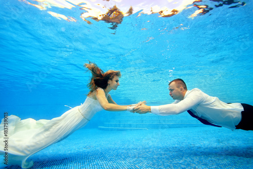 The bride and groom in wedding dresses swim underwater in the pool to meet each other. Portrait. Landscape orientation. Shooting under water