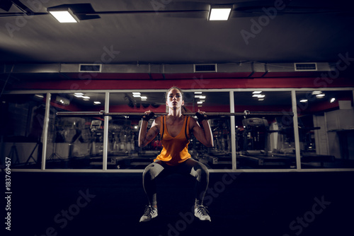 Close up portrait of motivated young shape active fitness woman crouch and doing squad exercise with a bar behind the neck in the gym and looking at the camera.