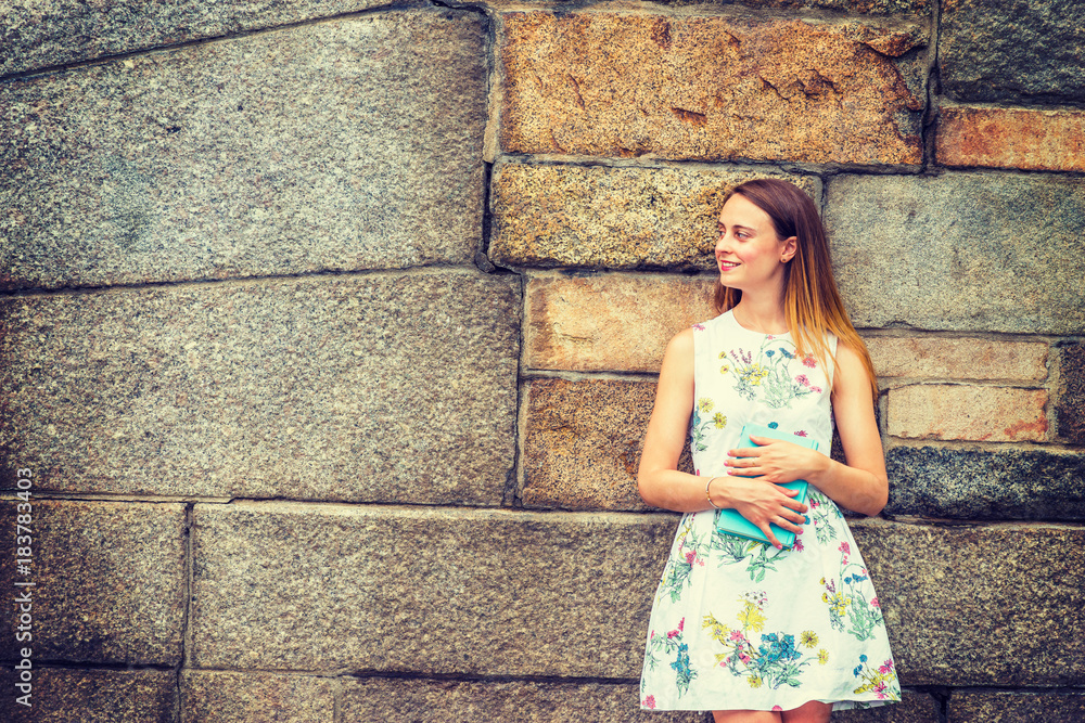 Portrait of College Student. A girl wearing flower patterned, sleeveless, white dress, holding green book, standing against rocky wall on campus, smiling, looking away.  Copy Space..