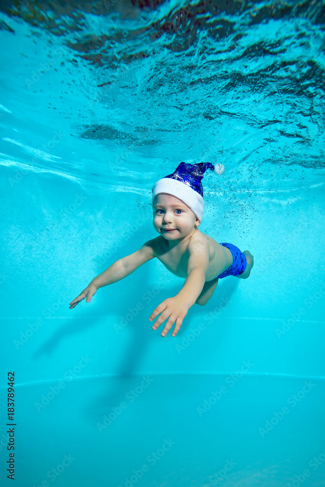 Adorable baby in hat of Santa Claus, with blue eyes swims underwater in the pool and looking at me. Portrait. Shooting under water. Vertical orientation