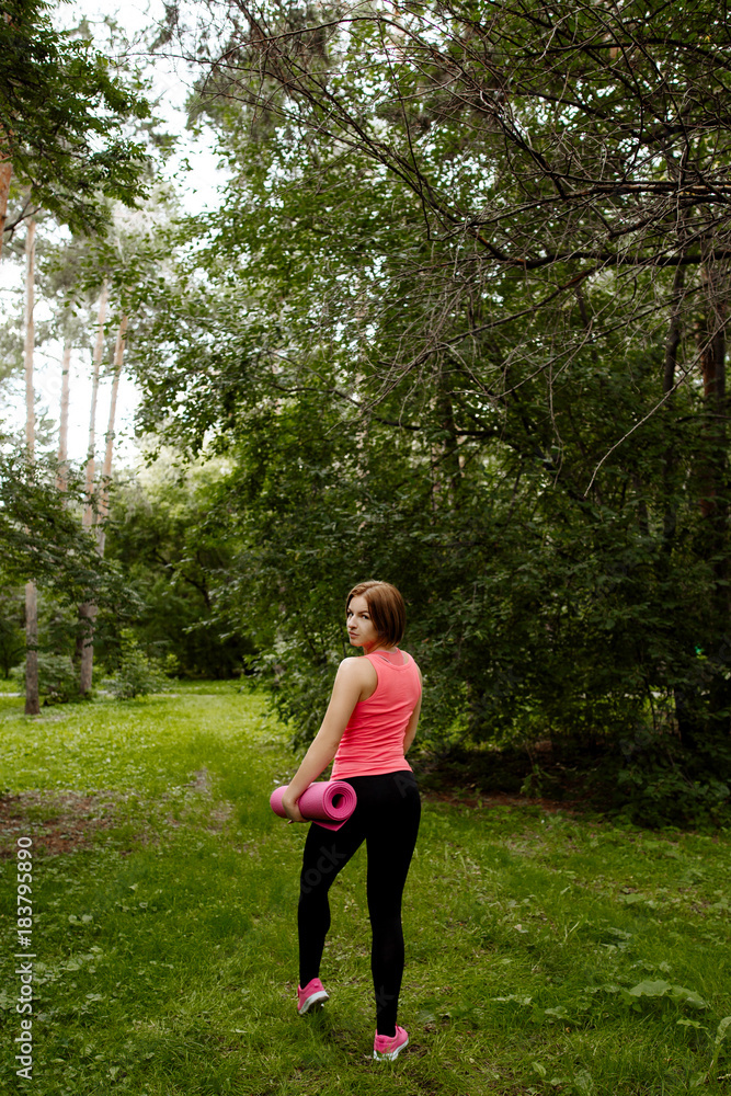 fitness in the fresh air. The sports girl in a pink undershirt, black leggings and pink sneakers against the background of green foliage. The woman does warm-up and charging on a rug for yoga. fitness in the fresh air. The sports girl in a pink undershirt, black leggings and pink sneakers against the background of green foliage. The woman does warm-up and charging on a rug for yoga.