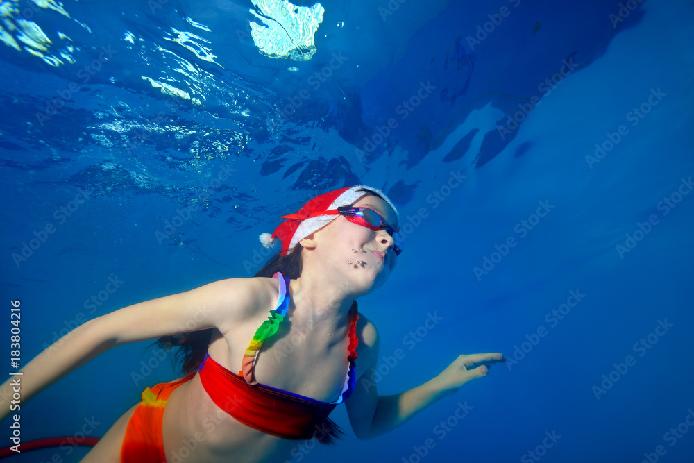 Little girl in hat of Santa Claus plays sports and swims underwater in the pool. Portrait. Shooting under water. Horizontal orientation