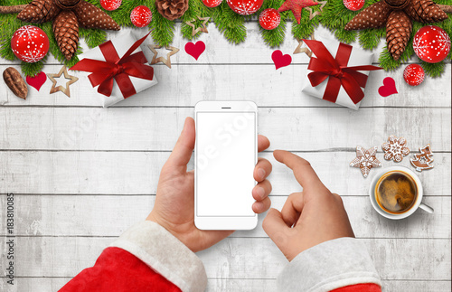 Santa's hand holding and touching white smartphone with empty screen at bright wooden desk with Christmas decoration, gift boxes, coffee and cakes. Top view
