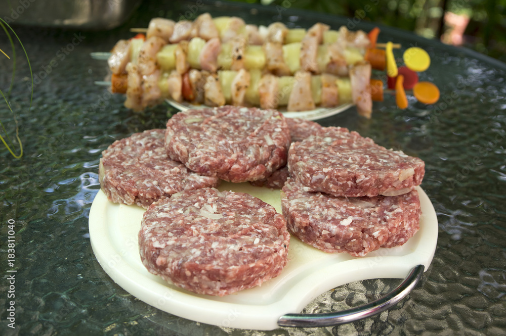 Raw pork and beef meat prepared for hamburgers on white rounded cutting board, chicken skewers on background, ready for grill
