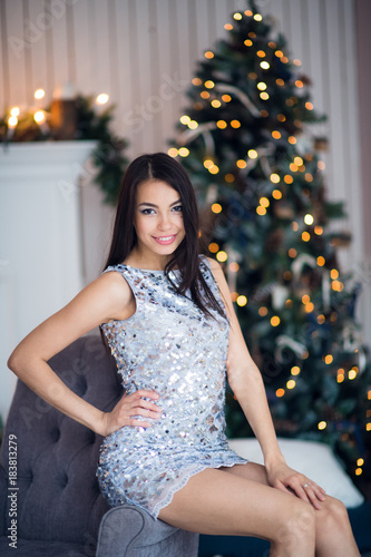 Christmas. Cute girl wearing shiny dress at home sitting on armchair against fireplace and fir tree