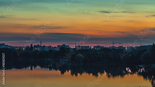 Beautiful sunset skyline of a city and trees with a lake in the foreground © Artsiom