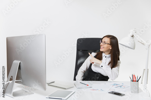 Smiling business woman sitting at the desk, working at computer with documents in light office, pointing her finger at white background with copy space for your advertisement or promotional content