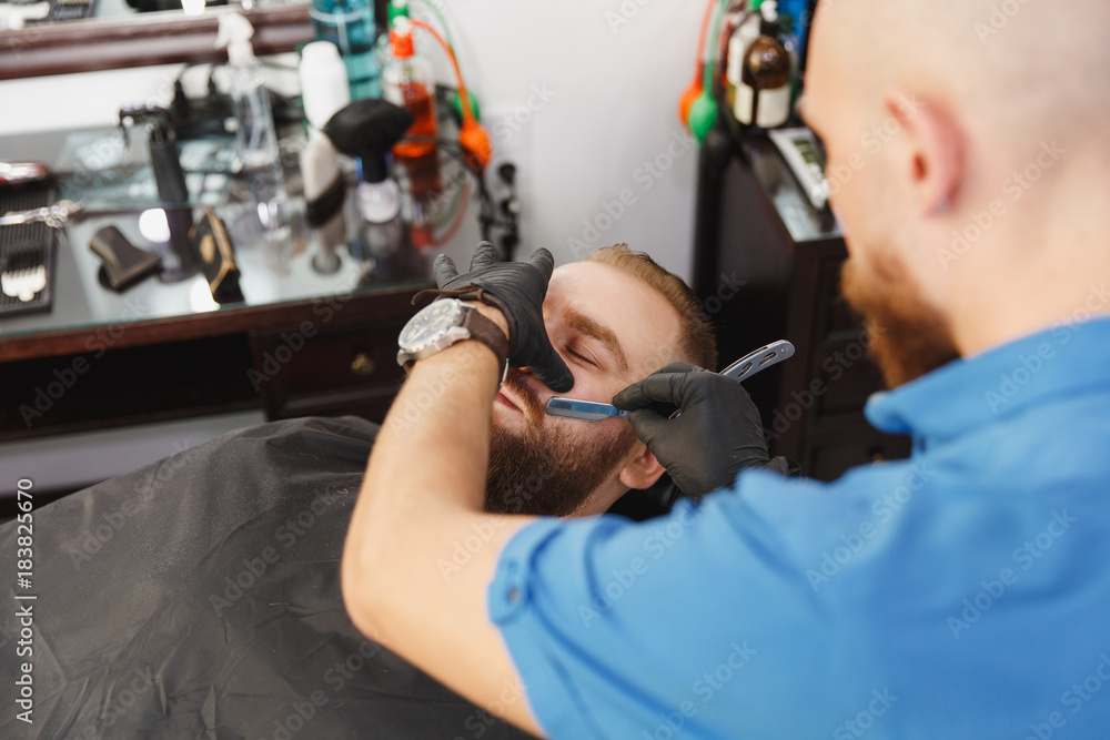 Male professional hairdresser serving client, shaving thick big beard straight razor. Ginger handsome stylish young man with short hair getting trendy haircut, black cape. Light white barber shop room Male professional hairdresser serving client, shaving thick big beard straight razor. Ginger handsome stylish young man with short hair getting trendy haircut, black cape. Light white barber shop room