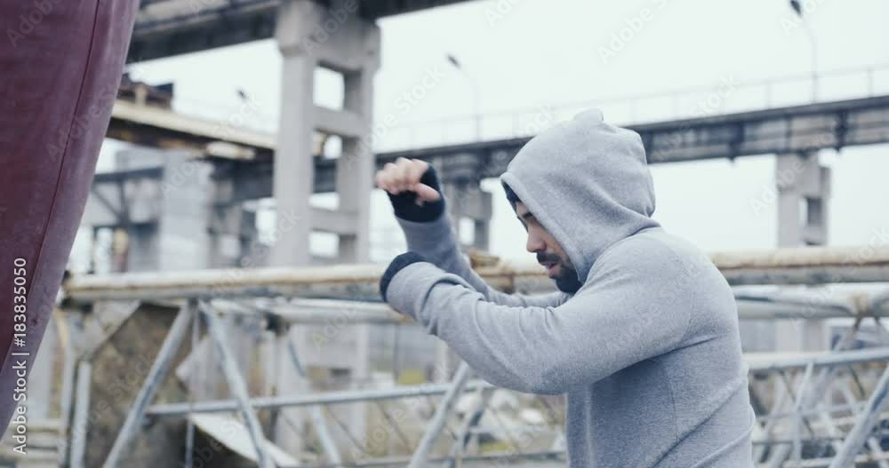 Attractive man with a beard in a sporty grey coat and hood kicking a bag on the thrown old factory on the cold misty day. Outdoor Attractive man with a beard in a sporty grey coat and hood kicking a bag on the thrown old factory on the cold misty day. Outdoor