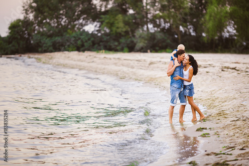 Young beautiful and stylishly dressed family of three people mom, dad and daughter of one year on the shoulders of a man walking along the seashore barefoot holding his hand