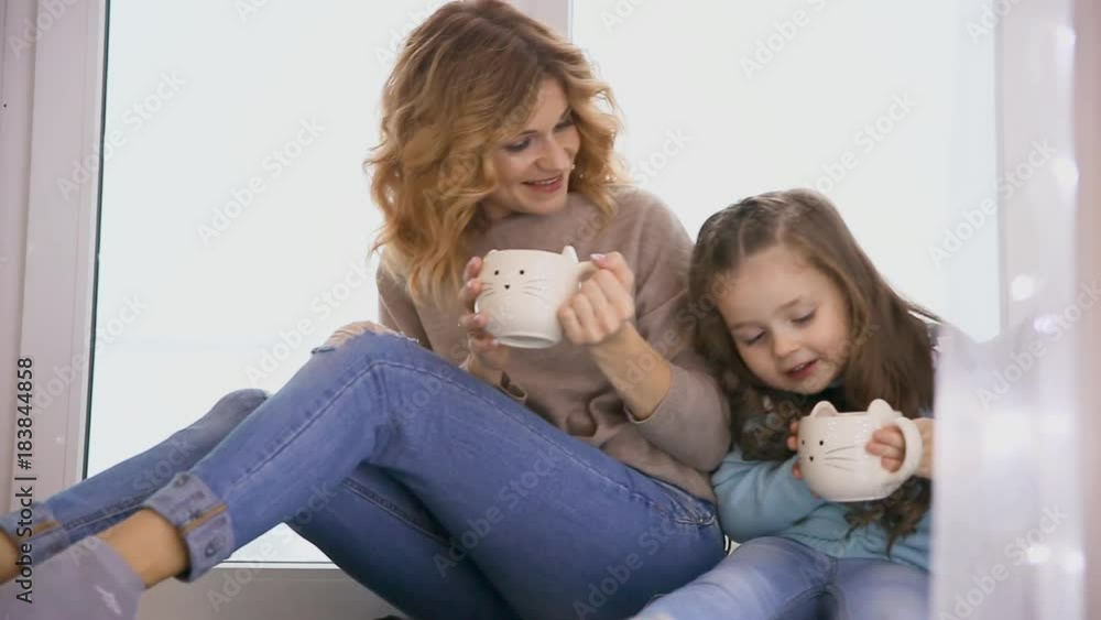 Mother and daughter sitting on the window sill in sweaters at Christmas and looking out the window, talk to each other and drink coffee. Mother and daughter sitting on the window sill in sweaters at Christmas and looking out the window, talk to each other and drink coffee.
