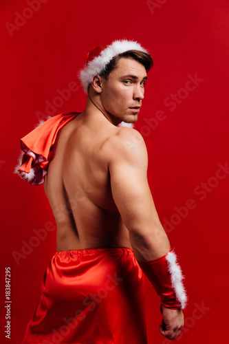 christmas holidays. sexy strong santa claus wearing hat. Young muscular man. red background.