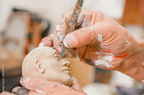 Close up of man ceramist hands holding a tool and working on sculpture details of the head on wooden table in workshop