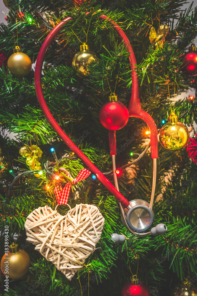 Close up view of a red stethoscope hanging on a decorated Christmas tree with baubles or balls, hearts and shining colorful Christmas lights. Medical Christmas or Holiday in hospital concept