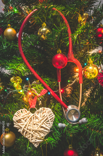 Close up view of a red stethoscope hanging on a decorated Christmas tree with baubles or balls, hearts and shining colorful Christmas lights. Medical Christmas or Holiday in hospital concept photo