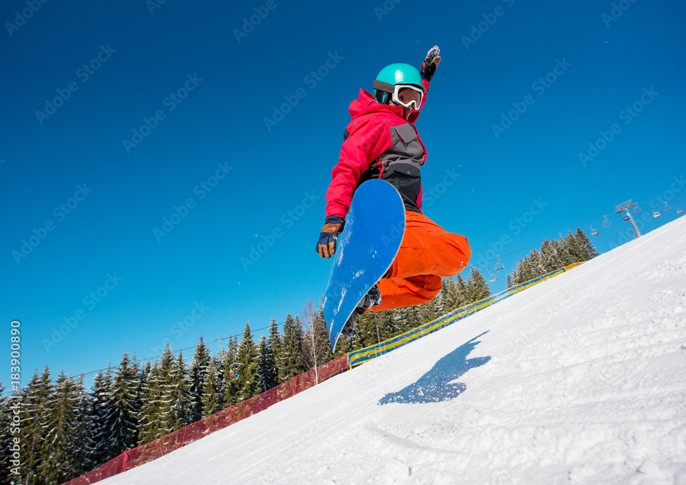 Low angle shot of a snowboarder jumping in the air while riding on the slope in the mountains on a beautiful sunny winter day. Forest, blue sky and ski-lift on the background
