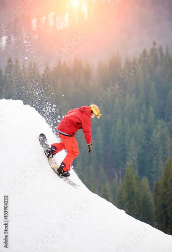 Male snowboarder freerider in a red suit riding from the top of the snowy hill with snowboard in the evening at sunset. Skiing and snowboarding concept