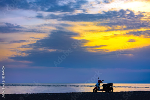 Fototapeta Naklejka Na Ścianę i Meble -  Scooter silhouette on a dark bank in front of the sea shore against the background of a beautiful orange sky with blue clouds. Sunset on the Black Sea, Adler, Sochi, Russia.