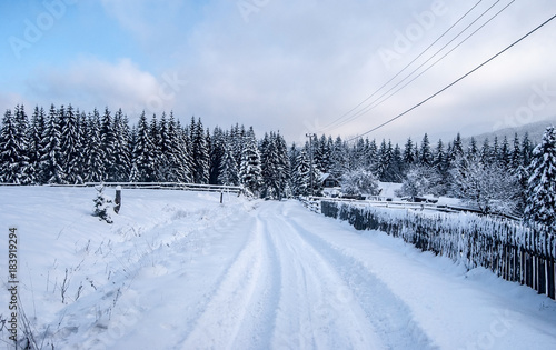 winter countryside with snow covered road, isolated house with fence, forest and blue sky with clouds