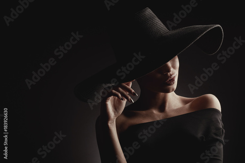 Dramatic dark studio portrait of elegant woman in black wide hat and black dress. Hidden eyes.