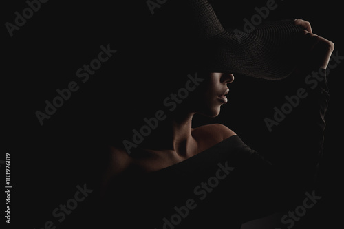 Dramatic dark studio portrait of elegant and luxury woman in black wide hat and black dress. Hidden eyes.