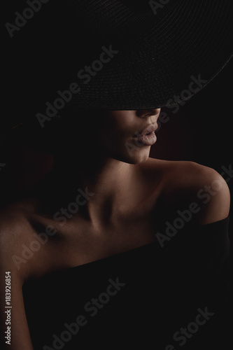 Dramatic dark studio portrait of elegant and luxury woman with beautiful hands, wearing black wide hat and black dress. Hidden eyes.