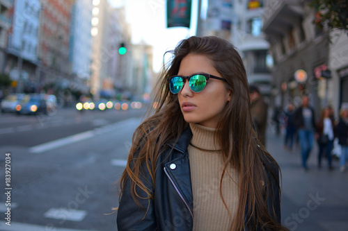 Mujer  joven estilosa  con gafas de sol y chaqueta negra caminando o realizando compras por la calle gran via de la ciudad de Madrid durante el atardecer