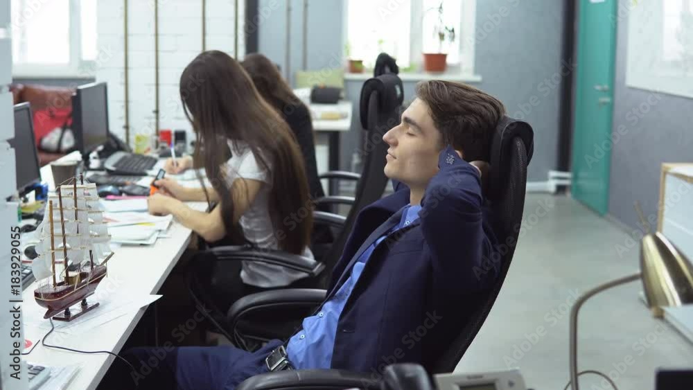 Weekend is close. Young positive man in suit is sitting in the workplace and dreaming of vacation with his eyes closed against the background of working female employees Weekend is close. Young positive man in suit is sitting in the workplace and dreaming of vacation with his eyes closed against the background of working female employees