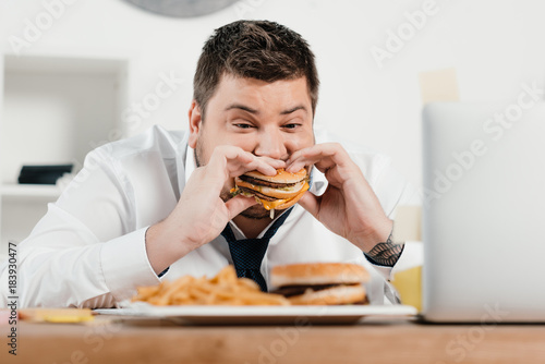 overweight businessman eating hamburger and french fries in office
