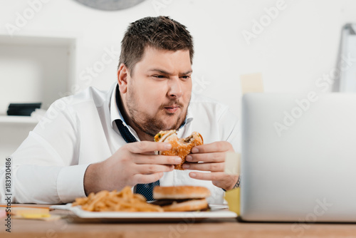 Bild auf Leinwand overweight businessman working while eating hamburger and french fries in office