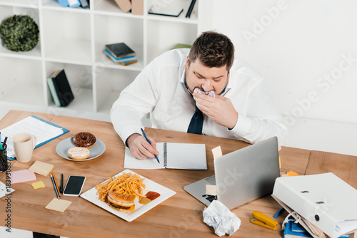 overweight businessman eating donuts, hamburger and french fries while wokring in office photo