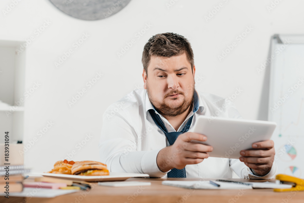 overweight businessman looking at junk food while working with tablet at workplace