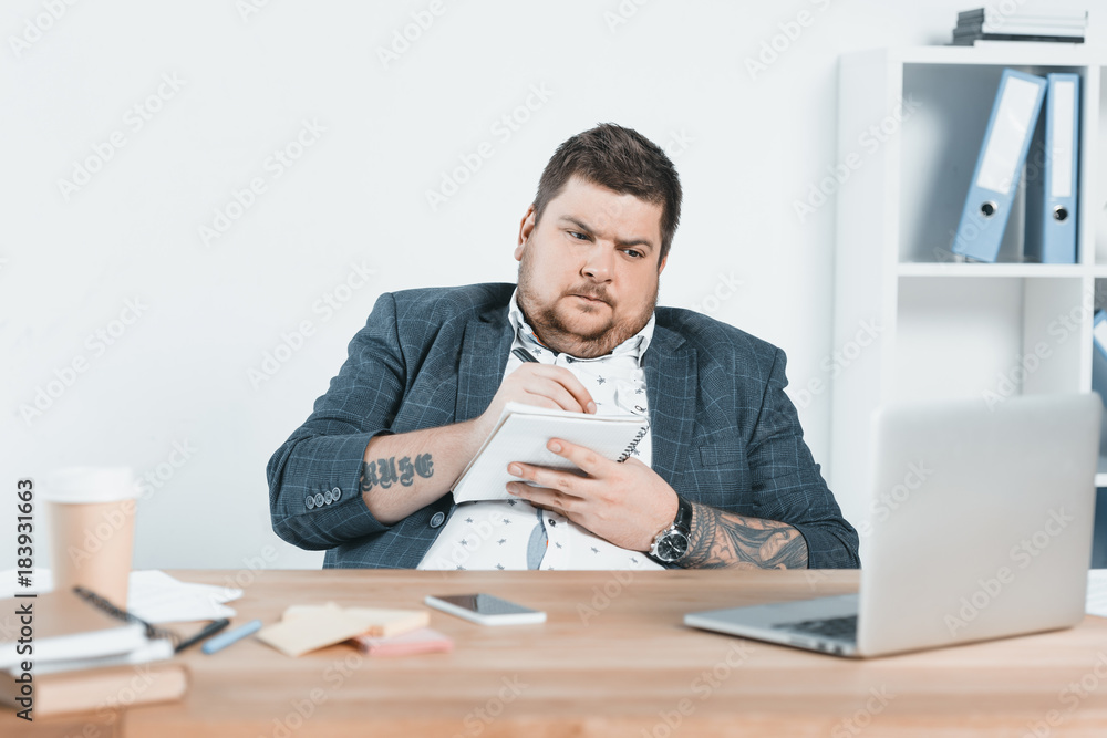 overweight businessman in suit writing in notepad and working with laptop at workplace overweight businessman in suit writing in notepad and working with laptop at workplace