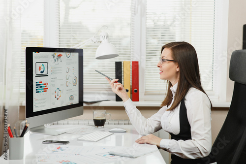 Beautiful business woman in suit and glasses working at computer with documents in light office, looking and pointing at the monitor © ViDi Studio